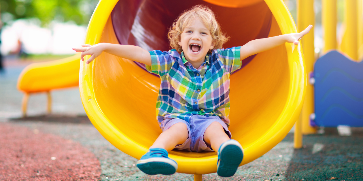 A little boy smiling with arms out at the bottom of a yellow slide
